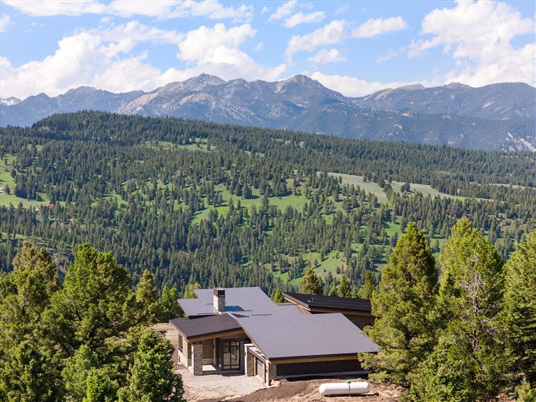 Panoramic view of a mountain valley with evergreen trees, a lake, and a house under a clear sky.